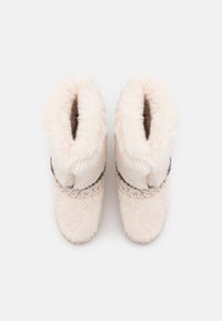 Botas peludas de color crema con una textura suave, que presentan una banda decorativa trenzada y herrajes plateados alrededor del tobillo.