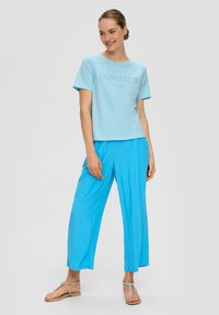 Camiseta de algodón azul claro con la palabra "VERANO" impresa en un diseño texturizado, combinada con pantalones sueltos de un azul a juego. Se lleva con sandalias minimalistas.