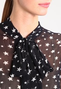 Svart genomskinlig blus med en fjärilsknyt vid halsringningen, prydd med vita stjärnmönster över hela. Mjuk, lättviktsmaterial.