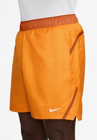 Orangene Sportshorts mit einem strukturierten Rautenmuster, elastischen Bund und kontrastierenden braunen Akzenten. Mit einem weißen Nike-Logo.