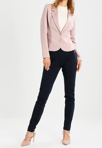 Lichtroze blazer met twee knopen, getailleerd ontwerp; gecombineerd met donkerblauwe skinny broek. Beige hoge hakken maken de outfit compleet.