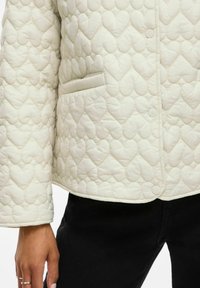 Gequilted jack in gebroken wit met hartvormige patronen, voorzien van een enkele knoopsluiting en een zijzak. Zachte textuur, losse pasvorm.