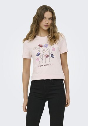 Giovane donna con una t-shirt rosa chiaro con fiori colorati e il testo "BLOOM AS YOU ARE,", abbinata a pantaloni neri, in piedi su uno sfondo semplice.