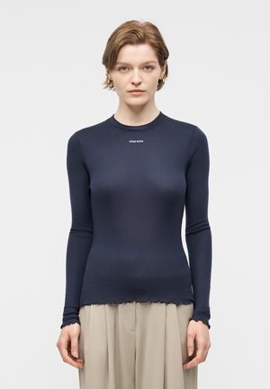 Νεαρή γυναίκα με κοντά μαλλιά, που φοράει στενή μπλε σκούρα μπλούζα με μακριά μανίκια και το κείμενο «STINE GOYA» και μπεζ παντελόνι με φαρδιά πόδια, στέκεται μπροστά σε λευκό φόντο.