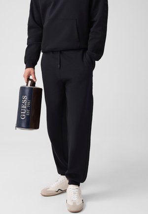 Person trägt schwarzen Pullover und Hose, hält eine dunkle GUESS-Tasche und trägt weiße Sneaker mit beigen Sohlen, steht vor hellem Hintergrund.