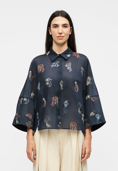 Femme portant un chemisier transparent bleu marine à boutons, aux manches larges et aux imprimés abstraits colorés, associé à un pantalon plissé beige taille haute.