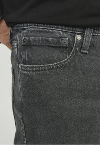 Jeans de mezclilla negra con un acabado desteñido, que presentan un bolsillo delantero con remaches de metal, detalles de costura sutiles y una superficie texturizada.