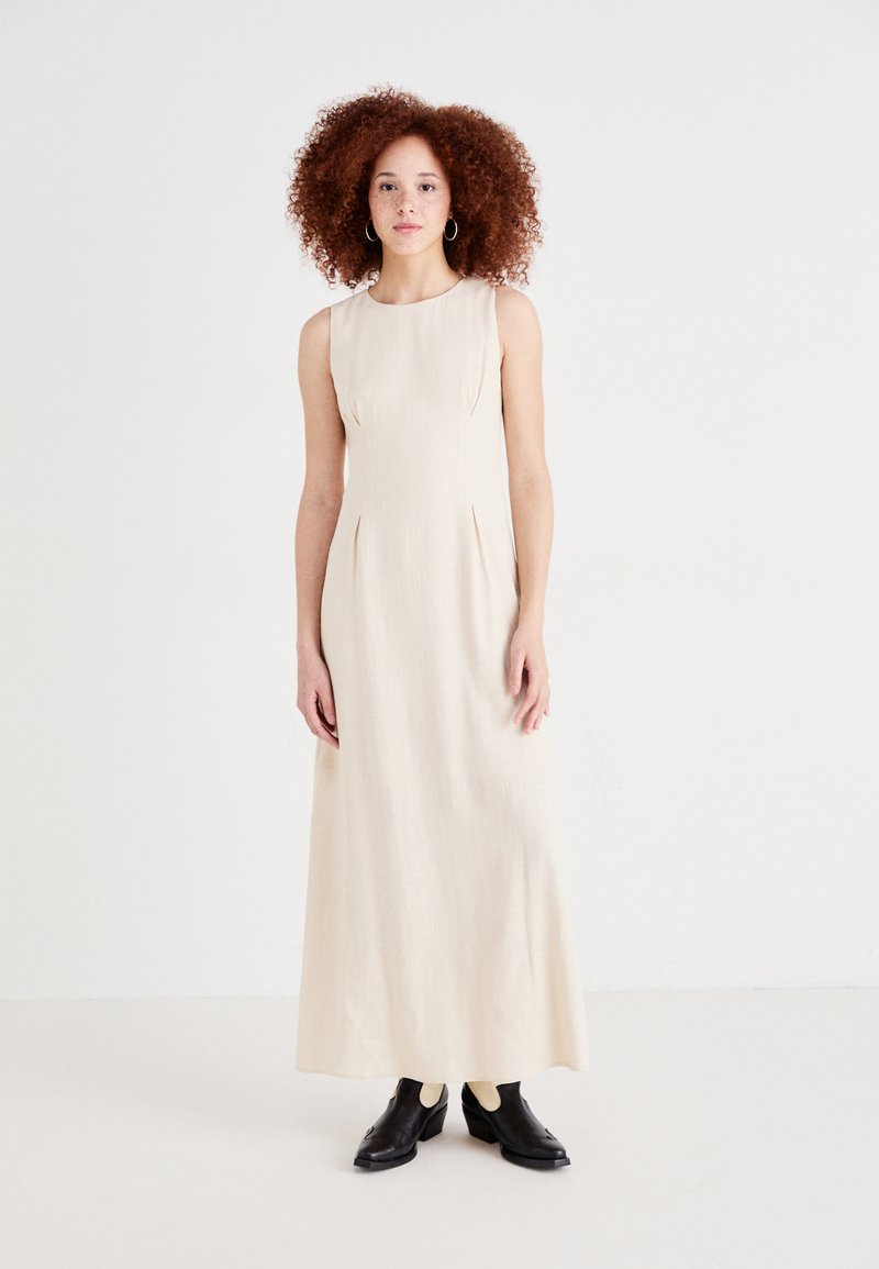 Robe maxi sans manches beige clair avec une coupe structurée, ornée de légers plis, accompagnée de chaussures noires texturées.