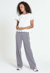 Top bianco a maniche corte con bordi ondulati abbinato a pantaloni grigi a gamba larga; modello indossa scarpe sportive con dettagli rossi.