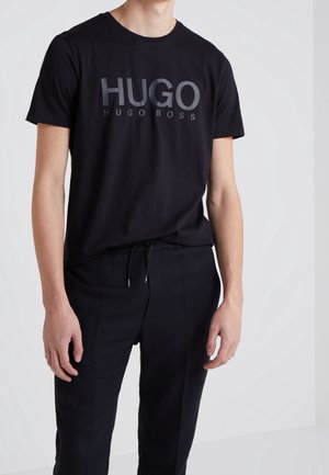 Mężczyzna w czarnej koszulce Hugo Boss i czarnych spodniach ze sznurkiem na gładkim jasnym tle.