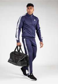 Marineblauwe tracksuit met witte logo-accenten, ritsjack en bijpassende broek. Zwarte sporttas in de hand, gecombineerd met zwarte sportschoenen.