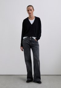 Jonge vrouw staat tegen een grijze muur en draagt een zwarte V-hals trui over een wit shirt, grijze wijde jeans en zwarte schoenen.