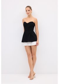 Černé šaty bez ramínek s peplum střihem, fitted korzetem a vrstvenou bílou plisovanou sukní. Modelka má na sobě zlaté náušnice ve tvaru kruhu a béžové sandály na tenkých páscích.
