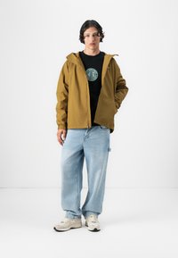 Brun letvægtsjakke med hætte, lynlås, elastiske manchetter, båret over en sort T-shirt med et rundt grønt logo; parret med lysblå jeans.