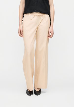 Person iført beige bukser med vide ben og bindebånd foran samt sorte spidse sko, kombineret med en sort ærmeløs top.