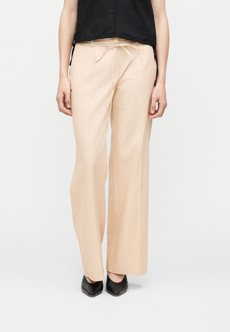 Άτομο που φοράει μπεζ παντελόνι με φαρδιά μπατζάκια και μπροστινό δέσιμο, μαζί με μαύρα παπούτσια με μυτερή μύτη και μαύρο αμάνικο τοπ.