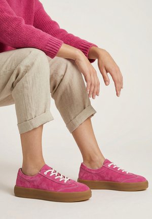 Osoba nosząca jaskrawo różowe trampki z białymi sznurówkami, beżowe podwinięte spodnie i pasujący różowy sweter, siedząca ze zrelaksowanymi rękami.