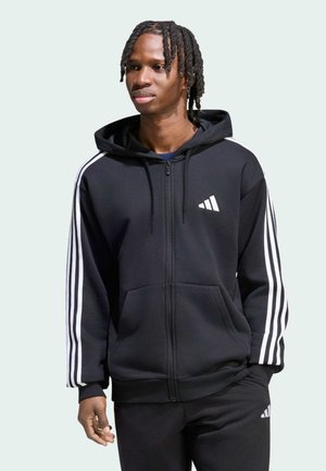 Junger Mann mit geflochtenem Haar, der einen schwarzen Adidas Zip-Hoodie und passende Hosen mit weißen Streifen an Ärmeln und Beinen trägt.