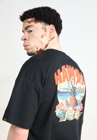 Černé tričko s barevným potiskem ananasu s tropickými květinami a textem "HAWAII Aloha" na zadní straně. Krátké rukávy.