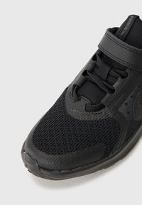 Chaussure de sport noire avec un dessus en maille, accents texturés et semelle en caoutchouc moulé. Comprend des œillets pour les lacets et une sangle sur le coup de pied.