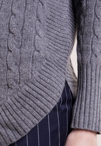 Suéter gris de punto con un patrón de trenzas, puños acanalados y un dobladillo más corto en la parte delantera. Textura suave contra pantalones Azul marino con rayas.