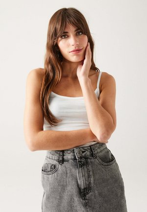 Jeune femme aux longs cheveux bruns, portant un débardeur blanc et un jean gris taille haute, reposant sa main sur son visage.