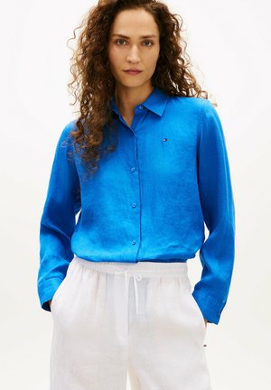 Kvinde med krøllet hår iført en lys blå skjorte med knapper og løse hvide bukser, stående med hænderne i lommerne mod en lys baggrund.