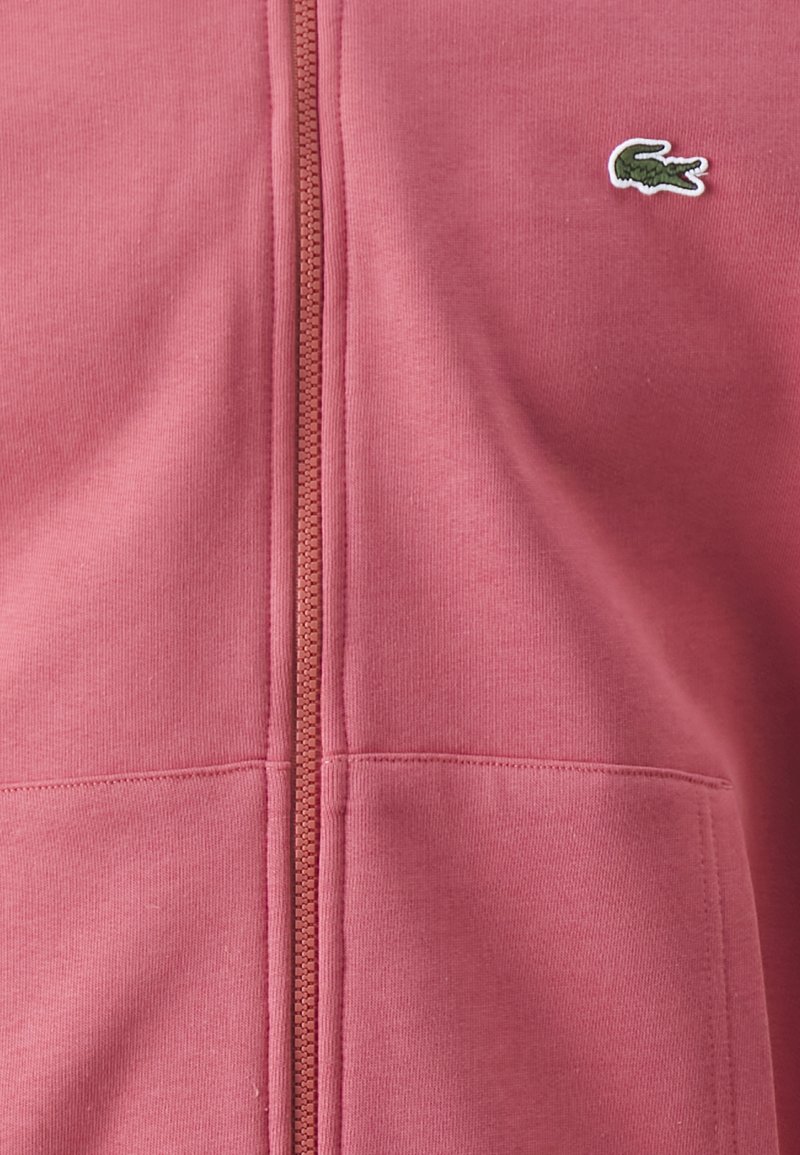 Haut à capuche zippé rose en tissu doux, avec une surface texturée, une fermeture éclair sur le devant et un petit logo brodé vert sur la poitrine.