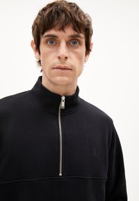 Sweat-shirt zippé noir avec col haut, texture douce et détail de logo subtil sur la poitrine. Accent de fermeture éclair argentée.