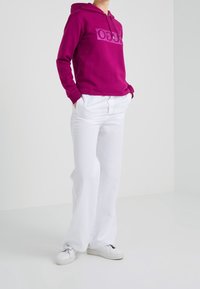 Sweat à capuche magenta avec un logo sur le devant, associé à un pantalon large blanc et des baskets blanches. Le tissu semble doux et décontracté.