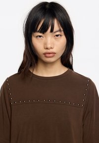 Camiseta marrón de manga corta con una textura suave y adornos de tachuelas metálicas en los hombros y el área del pecho. Cuello redondo.