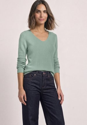 Femme portant un pull vert clair à col en V et un jean bleu foncé, debout devant un fond clair uni.