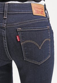 Jeans de mezclilla azul oscuro con costuras amarillas contrastantes, que presentan una etiqueta de cuero con la inscripción "Levi Strauss & Co." y una etiqueta roja en el lateral.