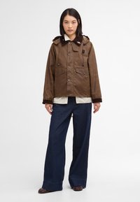 Brown waxed jacket with a hood, snap buttons, and two front pockets. Paired with a striped shirt and wide-leg dark blue jeans.