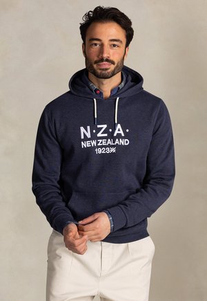 Man draagt een marineblauwe hoodie met de tekst "N.Z.A. New Zealand 1923", beige broek en een geruit overhemd eronder, staand tegen een effen achtergrond.
