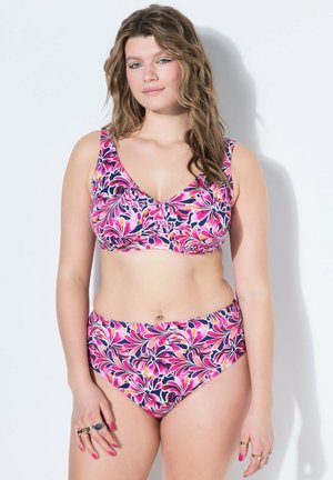 Frau mit welligem Haar, die einen rosa, lila und weißen Blumenbikini trägt und mit neutralem Gesichtsausdruck vor einer weißen Wand steht.