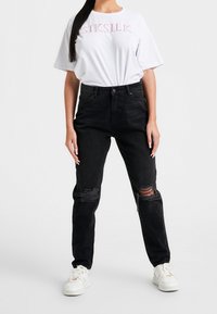 Černé džínové kalhoty s roztržením na kolenou, spárované s bílým tričkem s krátkým rukávem, které má růžový nápis. Bílí tenisky dokreslují celkový vzhled.