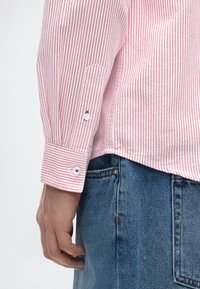 Ριγέ πουκάμισο μακρυμάνικο σε ανοιχτό κόκκινο και λευκό με κουμπιά στους μανσέτες, συνδυασμένο με μπλε τζιν τζιν. Ορατές λεπτομέρειες περιλαμβάνουν κουμπιά και ραφές.