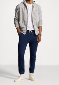 Felpa grigia chiara con zip e tasche, t-shirt bianca sotto, pantaloni joggers blu scuro con coulisse e scarpe beige con suole texture.