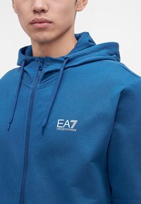 Мъж, носещ син суичър с качулка и връзки, с бял лого EA7 Emporio Armani на гърдите.
