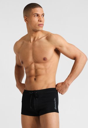 Black swim shorts with a drawstring, featuring a logo on the left side. The material is smooth with a fitted design and contrasting stitching.