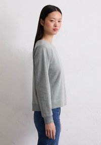 Sudadera gris claro con cuello redondo y mangas largas, que presenta puños acanalados y un corte relajado. Combinada con vaqueros azul oscuro.
