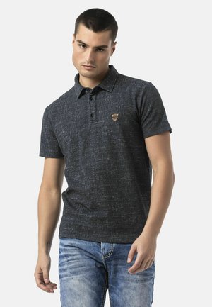 Jonge man draagt een donker textuur korte mouw polo en lichtblauwe vervaagde spijkerbroek, staand tegen een effen achtergrond.