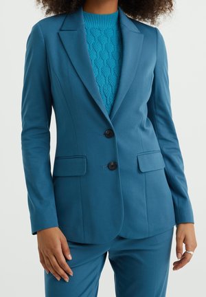 Kobieta ubrana w turkusową marynarkę z dwoma guzikami i dopasowane spodnie, w połączeniu z teksturowanym, niebieskim swetrem dzianinowym pod spodem.