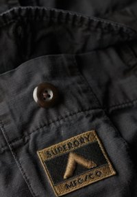 Zwarte stof met een ronde bruine knoop en een rechthoekig geweven label met de tekst "Superdry" en een driehoekig logo in goud en zwart.