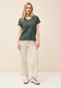 Top verde a maniche corte con texture e scollatura a V, abbinato a pantaloni a gamba larga color crema e sneakers bianche con dettagli rossi.