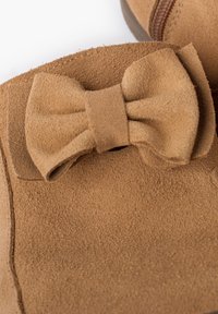 Botas de tobillo de ante color tostado con un suave lazo decorativo en un lado. La textura es lisa y suave, con un cierre de cremallera a lo largo de un lado.