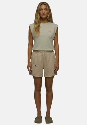 Femme aux longs cheveux portant un haut beige sans manches, un short beige assorti avec des poches et des chaussures marron à enfiler, debout devant un fond uni.