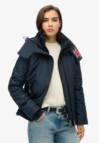 Granatowa kurtka puchowa z kapturem, z zapięciem na suwak, prążkowanymi mankietami i naszywką z logo na ramieniu; zestawiona z kremowym swetrem i dżinsami.