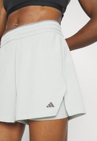 Pantaloni sportivi verde chiaro con un motivo testurizzato, vita elastica e fessure laterali. Presenta un logo grigio Adidas in basso a sinistra.